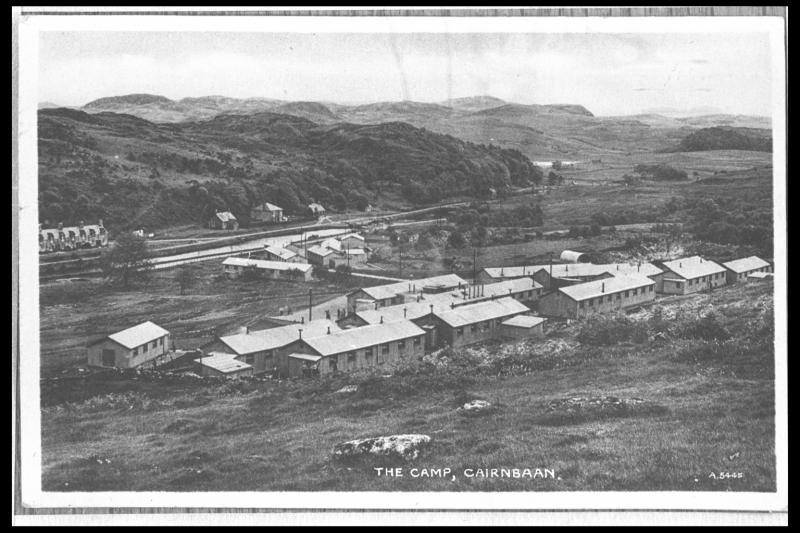 A black and white postcard showing a cluster of long and low rectangular camp buildings set within an upland landscape of rough ground and rock outcrops